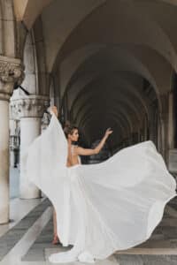 Eine Frau in einem fließenden weißen Kleid, das an eine Braut an ihrem Hochzeitstag erinnert, posiert anmutig unter einem eleganten Steinbogen. Das Kleid bauscht sich dramatisch und erzeugt vor dem Hintergrund symmetrisch angeordneter Säulen und Bögen ein Gefühl von Bewegung.
