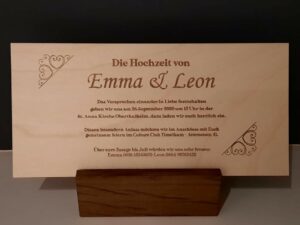 Eine hölzerne Hochzeitseinladungskarte mit dekorativen Herzmustern, in deutscher Sprache, lädt die Gäste zur Hochzeit von Emma und Leon am 26. September 2020 in der St. Anna Kirche Oberthalheim mit anschließender Feier im Kulturclub Timelkam ein.