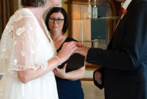 Braut und Bräutigam tauschen während ihrer Hochzeit Ringe aus. Die Braut, die ein weißes Kleid mit Blumendetails trägt, hält die Hand des Bräutigams. Eine Frau in einem marineblauen Kleid steht im Hintergrund und leitet die Zeremonie, wodurch jeder Moment dieser Hochzeit unvergesslich wird.