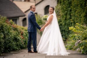 Eine Braut in weißem Kleid und Schleier hält einen Blumenstrauß und steht mit dem Bräutigam im blauen Anzug auf einem Steinweg. Sie lächeln in die Kamera und feiern ihre Hochzeit, umgeben von üppigem Grün und charmanten Gebäuden im Hintergrund.