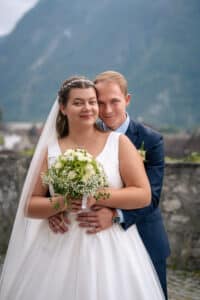 Vor einer malerischen Bergkulisse umarmen sich Braut und Bräutigam glücklich im Freien. Während die Braut einen Strauß weißer Blumen umklammert, legt der Bräutigam seine Arme um ihre Taille und verkörpert die Freude des Heiratens in dieser idyllischen Umgebung.