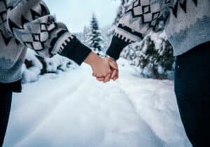 Zwei Menschen halten sich in einer verschneiten Landschaft an den Händen, was an eine romantische Winterhochzeit erinnert. Beide tragen kuschelige, gemusterte Pullover. Schneebedeckte Bäume und ein schneebedeckter Weg schaffen eine friedliche, winterliche Szene, die perfekt für Heiratsträume ist.