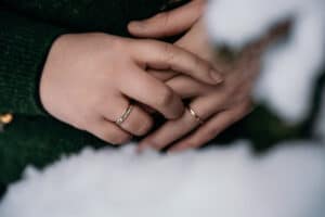Nahaufnahme einer Person, die die Hände ineinander verschränkt hat und an jeder Hand einen Ring trägt – ein Symbol für Heiraten. Die Person trägt einen dunkelgrünen Pullover und im Vordergrund liegt Schnee, was die heitere Schönheit dieses Augenblicks noch verstärkt.