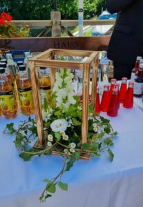 Eine hölzerne Laterne mit weißen Blumen und Grünzeug schmückt den Tisch und passt perfekt zu jeder Hochzeitssituation. Umgeben von klaren und roten Flaschen mit verschiedenen Getränken steht sie vor einer sonnigen Außenkulisse mit einem Holzgeländer und üppigem Laub – ideal für all Ihre Hochzeitsplanungsträume.