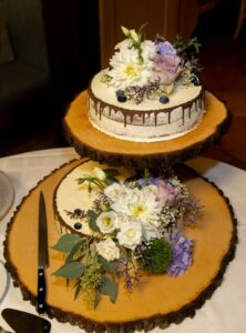 Eine zweistöckige rustikale Hochzeitstorte steht auf Holzplatten und ist mit weißen und violetten Blumen, Grünpflanzen und Blaubeeren geschmückt. Neben der Torte liegt ein Messer, das das romantische Hochzeitsthema perfekt ergänzt.