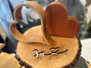 Zwei Holzherzen, eines hohl und eines massiv, ruhen auf einer rustikalen Scheibe. Das Wort „Ja“ mit Herz ist elegant auf die Oberfläche gemalt und fängt den Geist von Heiraten in dieser gemütlichen Umgebung perfekt ein.