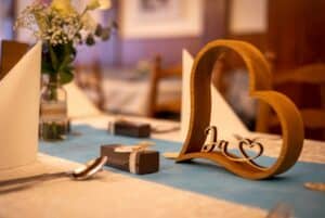 Eine hölzerne Herzdekoration mit dem Wort „Ja“ ziert einen Esstisch, perfekt für eine Hochzeit. Daneben liegen verpackte Geschenke, gefaltete weiße Servietten und eine kleine Blumenvase auf einem blauen Tischläufer. Der Hintergrund ist sanft verschwommen und schafft eine warme, einladende Atmosphäre, ideal für die Hochzeitsplanung.