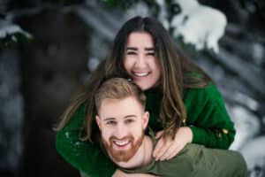 Eine lächelnde Frau in einem grünen Pullover sitzt spielerisch huckepack auf einem lächelnden Mann mit Bart in einer verschneiten Außenkulisse und fängt die Freude ihrer Winterhochzeit ein. Beide schwelgen in der winterlichen Atmosphäre, wobei schneebedeckte Bäume die zauberhafte Kulisse ergänzen.