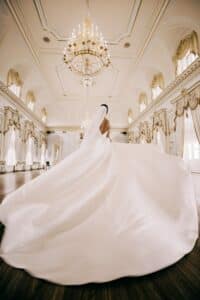 Eine Braut in einem voluminösen weißen Kleid steht in einem eleganten Ballsaal und verkörpert die Essenz einer Hochzeit. Tageslicht fällt durch große, drapierte Fenster, wirft einen anmutigen Schein und verstärkt die romantische Atmosphäre – eine exquisite Szene für alle, die Hochzeitsplanung betreiben.