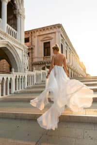 Eine Frau in einem fließenden weißen Kleid steigt bei Sonnenuntergang die Steintreppe neben einem kunstvollen, historischen Gebäude hinauf und verkörpert die Essenz einer romantischen Hochzeit. Das Kleid bauscht sich anmutig und fängt das sanfte Licht ein, während die Architektur ein klassisches europäisches Ambiente bietet, das perfekt für eine Hochzeit ist.