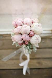 Ein Strauß hellrosa Pfingstrosen, perfekt für eine Hochzeit, ist mit einem weißen Band zusammengebunden. Die Blumen stehen vor einem rustikalen Holzhintergrund und schaffen eine sanfte und romantische Atmosphäre, ideal für Ihre Hochzeitsplanung.