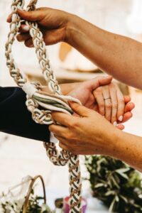 Eine Nahaufnahme von zwei Personen, die sich während einer Handfasting-Zeremonie bei ihrer Hochzeit an den Händen halten. Eine Person wickelt eine geflochtene Kordel um ihre verbundenen Hände, was Einheit und Verbundenheit symbolisiert. Grünpflanzen und ein unscharfer Hintergrund tragen zur intimen Atmosphäre bei, perfekt für diejenigen, die gerade mit der Hochzeitsplanung beschäftigt sind.
