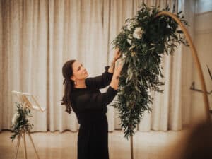 Eine Frau in einem schwarzen Kleid arrangiert Grünpflanzen und weiße Blumen auf einem Rundbogen im Innenbereich und verkörpert damit die Kunst der Hochzeitsplanung. Im Hintergrund sind ein heller Vorhang und ein kleines Blumenarrangement auf einem Ständer zu sehen, die die perfekte Kulisse für die bevorstehende Hochzeit schaffen.