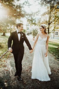 Ein Paar in Abendgarderobe, der Bräutigam im schwarzen Smoking und die Braut im weißen Kleid, gehen Hand in Hand einen sonnenbeschienenen, von Bäumen gesäumten Weg entlang. Während sie ihre Hochzeit planen, lächeln sie sich an, umgeben von einer herbstlichen Kulisse.