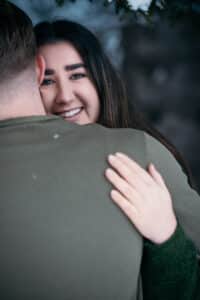 Eine Frau mit langem dunklem Haar lächelt, während sie eine Person in einem grünen Pullover umarmt und fängt einen zärtlichen Moment inmitten der Hektik der Hochzeitsplanung ein. Sie blickt über die Schulter der Person, ihre Hand ruht sanft auf ihrem Rücken, im Hintergrund sind die grünen Flecken verschwommen.