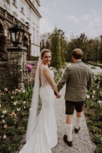 Eine Braut in weißem Kleid und Schleier hält die Hand eines Bräutigams in traditioneller Kleidung und geht einen Gartenweg entlang. Sie stehen draußen in der Nähe eines eleganten Gebäudes, umgeben von blühenden Blumen an einem sonnigen Hochzeitstag.