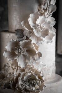 Eine Nahaufnahme einer elegant dekorierten mehrstöckigen Torte, perfekt für eine Hochzeit, mit aufwendigen weißen Zuckerblumen und zartem Blattwerk. Die Blumen haben detaillierte Blütenblätter und sind kunstvoll so angeordnet, dass sie über die Torte herabfallen, was ihr eine raffinierte Note verleiht und ideal für jede Hochzeitsplanung ist.