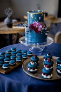 Eine mehrstöckige blaue Torte mit goldenen Akzenten und Blumen wird auf einem Glasständer präsentiert, perfekt für eine Traumhochzeit. Daneben ergänzen Tabletts mit blauen Macarons und kleinen Desserts, garniert mit Beeren und Sahne, das elegante Arrangement auf einer dunkelblauen Tischdecke.