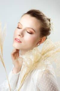 Eine Frau mit geschlossenen Augen, dezentem Make-up und zartem Haarschmuck posiert friedlich mit Pampasgras. Ihr strukturiertes weißes Outfit suggeriert Brauteleganz, perfekt für Hochzeitsträume vor einem schlichten, hellen Hintergrund, der eine verträumte, heitere Atmosphäre schafft.
