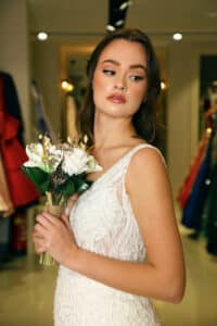 Eine Frau in einem weißen, strukturierten Kleid, perfekt für ihre bevorstehende Hochzeit, hält einen kleinen Strauß weißer und getrockneter Blumen. Sie blickt mit nachdenklichem Gesichtsausdruck zur Seite. Der Hintergrund zeigt verschwommene Kleidungsstücke und warmes Licht und schafft so die ideale Szene für ihre Hochzeitsplanung.