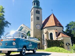 Ein blauer Oldtimer-Van von Volkswagen steht vor einer historischen Kirche und eignet sich perfekt als verträumte Hochzeitskulisse. Der hohe Glockenturm und das steile Dach verleihen der Szenerie Charme, während der klare Himmel und die Bäume die ideale Kulisse für Ihre Hochzeitsplanung an diesem sonnigen Tag bieten.