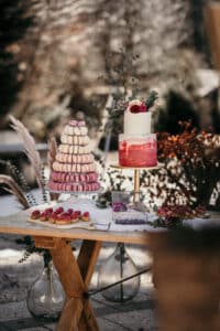 Ein Desserttisch, perfekt für eine Hochzeit, mit einem Macaron-Turm in Rosa- und Weißtönen, einer zweistöckigen Torte mit rot-weißem Zuckerguss, geschmückt mit Blumen, und einem Arrangement aus kleinem Gebäck und Süßigkeiten auf einem rustikalen Holztisch. Zur Dekoration gehört herbstliches Laub.