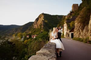 Braut und Bräutigam sitzen zusammen auf einer Steinmauer und blicken auf eine malerische Landschaft, perfekt für ihre Hochzeit. Sie sind umgeben von üppigen grünen Hügeln, ein paar Häusern und einer kurvenreichen Straße. Die Fernsicht zeigt ein Flussufer und ein altes Steingebäude auf der Hügelspitze, die ihre Traumkulisse für die Hochzeit verkörpern.