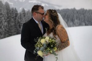 Inmitten einer verschneiten Landschaft umarmen sich Braut und Bräutigam an ihrem Hochzeitstag. Die Braut trägt ein weißes Kleid mit Schleier und hält einen Strauß weißer und gelber Blumen in der Hand. Ihr Tattoo verleiht der Szene Charme. Der Bräutigam, mit Brille und schwarzem Anzug mit beiger Krawatte, sieht aus, als wäre er bereit für ihre schöne gemeinsame Reise.