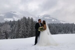 Braut und Bräutigam stehen auf einem schneebedeckten Feld, umgeben von immergrünen Bäumen und Bergen in der Ferne. Als Teil ihrer sorgfältigen Hochzeitsplanung hält die Braut einen Blumenstrauß in der Hand, während sie sich unter einem bewölkten Himmel anschauen. Ihre Kleidung bildet einen Kontrast zur ruhigen Winterlandschaft.