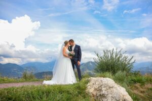 Braut und Bräutigam stehen mitten in ihrer Hochzeit eng beieinander auf einem grasbewachsenen Pfad mit einer Berglandschaft im Hintergrund. Die Braut trägt ein weißes Kleid und einen Schleier, während der Bräutigam einen dunklen Anzug trägt. Helle Wolken bedecken den Himmel, während sie diesen Hochzeitsmoment genießen.