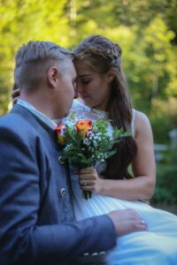 Ein Paar berührt sanft die Stirn, umgeben von üppigem Grün, und fängt die Essenz des Hochzeitsglücks ein. Die Frau im weißen Kleid hält einen Strauß Rosen und Schleierkraut, während der Mann einen Anzug mit dekorativen Knöpfen trägt. Die Szene ist friedlich und romantisch, perfekt für Hochzeiten.