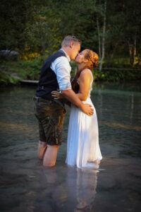 Ein Paar steht knietief in einem ruhigen, seichten Gewässer, umgeben von üppigem Grün. Der Mann in Unterhemd und Shorts küsst die Frau im weißen Kleid. Ihre romantische Umarmung deutet auf ihre Hochzeitspläne hin und hüllt die Szene in Träume von einer bezaubernden gemeinsamen Zukunft.