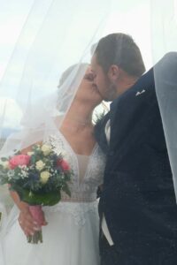 Braut und Bräutigam küssen sich bei ihrer Hochzeit unter einem hauchdünnen weißen Schleier. Die Braut hält einen Strauß rosafarbener und weißer Blumen in der Hand und trägt ein weißes Spitzenkleid. Der Bräutigam trägt einen dunklen Anzug mit einem beigen Einstecktuch. Die Hochzeitsszene ist romantisch und intim.