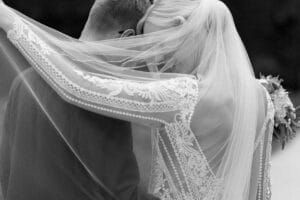 Ein Schwarzweißfoto fängt einen Hochzeitsmoment ein: Die Braut im Spitzenkleid und Schleier lehnt liebevoll an der Schulter des Bräutigams, ihr Schleier umhüllt sie beide teilweise und schafft so eine intime Erinnerung.