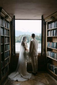 Braut und Bräutigam stehen Hand in Hand vor einem großen Fenster mit Blick auf einen ruhigen See und bewaldete Hügel. Während sie ihre Hochzeitsplanung planen, sind sie von Bücherregalen voller Bücher umgeben. Die Braut hält in ihrem langen Schleier sanft einen Blumenstrauß.