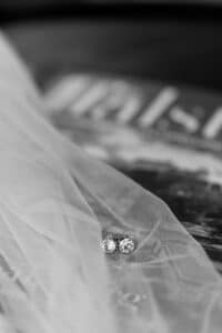 Schwarz-weißes Bild von zwei Diamantohrsteckern, die elegant auf einem transparenten Schleier ruhen und die Eleganz einer Hochzeit verkörpern. Der zarte Stoff bedeckt teilweise eine strukturierte Oberfläche oder ein bedrucktes Material im Hintergrund und verleiht dem Ganzen den perfekten Charme für eine Hochzeit.
