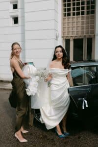 Eine Braut in einem weißen Kleid und Schleier, die einen Strauß weißer Blumen hält, steht an ihrem Hochzeitstag neben einem Auto. Eine Brautjungfer in einem grünen Kleid hilft beim Anziehen des Kleides. Die Szene spielt vor der Kulisse eines eleganten weißen Gebäudes mit hohen Fenstern, was diesem Moment des Hochzeitsglücks Charme verleiht.