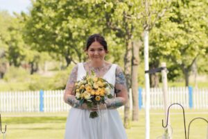 Eine Braut mit Tattoos lächelt, während sie einen Strauß gelber und weißer Blumen hält. Sie trägt ein ärmelloses weißes Kleid und steht draußen auf einer Rasenfläche mit Bäumen und einem weißen Zaun, was die Essenz ihrer Hochzeit wunderschön einfängt.