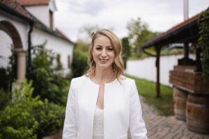 Eine Frau mit langen blonden Haaren und einem weißen Outfit steht lächelnd auf einem Kopfsteinpflasterweg und stellt sich vielleicht ihre Traumhochzeit vor. Im Hintergrund runden üppiges Grün und ein Gebäude mit einem roten Ziegeldach diese perfekte Szene für jede Hochzeitsplanung ab.