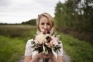 Eine Frau in einem Spitzenkleid steht im Freien und verkörpert die Essenz von Hochzeit, während sie einen bunten Blumenstrauß, darunter weiße und violette Blüten, nah an ihr Gesicht hält. Der Hintergrund zeigt eine verschwommene Landschaft mit Grün und einem bewölkten Himmel.