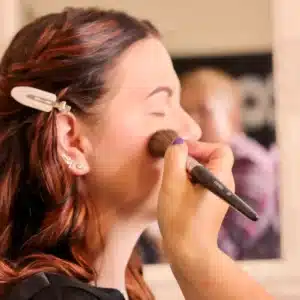Eine Person mit langen braunen Haaren lässt sich mit einem Pinsel Make-up auftragen, möglicherweise bereitet sie sich auf ihre Hochzeit vor. Ihre Augen sind geschlossen und eine Haarspange hält ihr Haar zurück. Der Hintergrund ist verschwommen, was die heitere Erwartung der Hochzeit noch verstärkt.