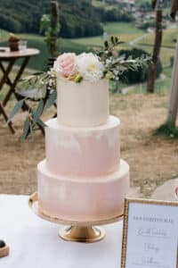 Eine dreistöckige rosa-weiße Hochzeitstorte mit Blumenschmuck steht auf einem goldenen Ständer im Freien und verkörpert die Eleganz einer perfekten Hochzeitsplanung. Im Hintergrund ist eine malerische Aussicht auf Bäume und Felder zu sehen, während ein gerahmtes Menü mit der Aufschrift „Hochzeitstorte“ die bezaubernde Atmosphäre ergänzt.