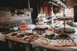 Ein rustikaler Hochzeits-Desserttisch im Freien mit dreistöckigen, mit Beeren dekorierten Torten, einem Turm aus Macarons, verschiedenen Gebäckstücken und Dessertgläsern. Holzwände und -balken umgeben die Szene und schaffen eine gemütliche, hüttenartige Atmosphäre, die perfekt für romantische Feiern ist.