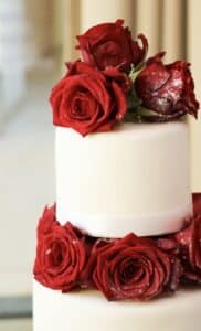 Eine zweistöckige weiße Torte, perfekt für eine Hochzeit, geschmückt mit leuchtend roten Rosen oben und um die Etagen herum, in einer sanft beleuchteten Umgebung.