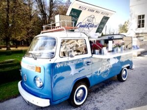 Ein blau-weißer Retro-Van mit „Coolkiste“-Branding wird in eine charmante mobile Bar verwandelt, perfekt für Ihre Hochzeitsplanung. Geschmückt mit Blumentöpfen und Flaschen auf der Theke steht sie im sonnigen Freien, umgeben von Bäumen und einem Gebäude, und schafft so eine idyllische Hochzeitskulisse.