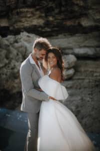 Ein Paar in Hochzeitskleidung steht eng aneinander gekuschelt an einem felsigen Strand und verkörpert die Essenz von Heiraten. Der Bräutigam trägt einen hellgrauen Anzug und die Braut sieht in einem schulterfreien weißen Kleid blendend aus. Sie lächeln herzlich in die Kamera und sind von großen Felsen umgeben, die von sanftem, natürlichem Licht beleuchtet werden.