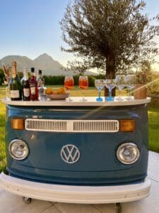 Ein VW-Camper verwandelt sich in eine charmante Bar, perfekt für jede Hochzeit, komplett mit Flaschen, Cocktails und Gläsern. Im Hintergrund entfaltet sich eine malerische Landschaft aus Bergen und Bäumen unter einem klaren Himmel und bietet eine idyllische Kulisse für Ihre Hochzeitsplanungsträume.