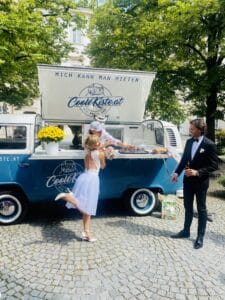 Eine Braut im weißen Kleid nimmt freudig Getränke von einem blauen Vintage-Foodtruck mit der Aufschrift „Coole Kiste.at“ bei ihrer Hochzeit entgegen. Ein Mann im schwarzen Anzug steht daneben, hält ein Getränk in der Hand und fängt den perfekten Moment der Hochzeitsmagie ein.