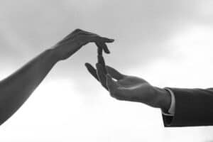 Zwei Hände, die sich vor einem hellen Hintergrund fast berühren, rufen die Vorfreude und Verbundenheit eines Hochzeitstages hervor. Eine Hand, die von einer Person im Anzug ausgestreckt wird, streckt sich mit leicht angewinkelten Fingern nach der anderen aus – und fängt die Essenz von Liebe und Engagement ein, die für die Hochzeitsplanung von zentraler Bedeutung sind.