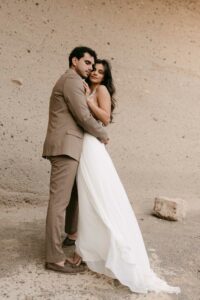 Ein Paar umarmt sich vor einer strukturierten, beigen Wand und verkörpert die Gelassenheit ihrer Hochzeit. Die Frau trägt ein fließendes weißes Kleid und der Mann einen braunen Anzug. Sie stehen eng beieinander auf einer rauen, sandigen Oberfläche und fangen einen Moment ein, der eine durchdachte Hochzeitsplanung verkörpert.